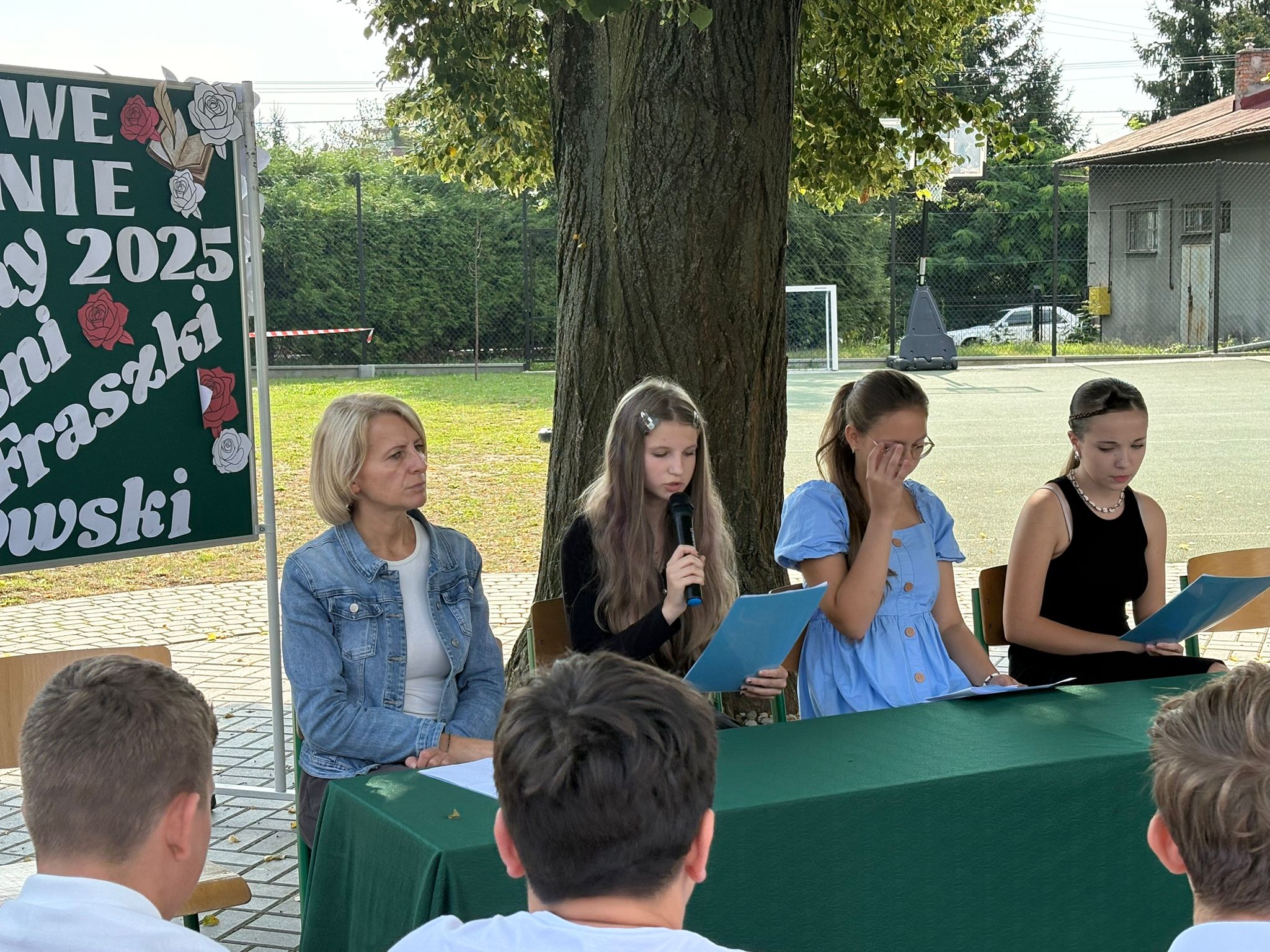Na zdjęciu widoczna jest tablica z napisem Narodowe Czytanie, widnieje wizerunek Jana Kochanowskiego. Przy stole siedzą uczniowie i Pani Dyrektor czytając pieśni, fraszki i treny.